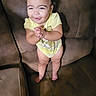 baby, child, toddler, standing, smiling, happy, face, portrait, indoors, living_room, couch, leather, furniture, yellow_shirt, onesie, barefoot, hands_clasped, dark_hair, candid, photography