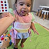 baby, child, toddler, standing, smiling, tongue_out, portrait, indoor, playroom, play_mat, toys, stuffed_animal, highchair, adult_hand, dress, striped_clothing, barefoot, sock, happy, face