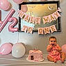 baby, balloon, banner, birthday, cake, celebration, cute, decorations, festive, happy, indoors, infant, mirror, party, pink, sitting, smiling, table, toddler, wall