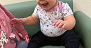 Penelope joined the competition — help win amazing prizes! baby, black_pants, chair, child, cute, earring, face, feet, floral_clothing, green_seat, hair, hands, happy, indoor, infant, pink_bag, portrait, sitting, smiling, toy