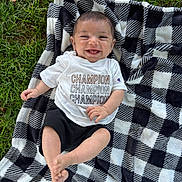 Billy is registered to the contest to win money with this photo: baby, smiling, child, blanket, checkered_pattern, grass, outdoor, shirt, clothing, shorts, happy, face, feet, hand, person, infant, lying_down, nature, casual, daylight