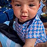 baby, child, face, portrait, blue_shirt, baseball_pattern, stadium, seats, people, hand, curious, close_up, infant, outdoor, daylight, casual, clothing, person, sitting, event