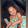 toddler, child, smiling, curly_hair, car_seat, blanket, cartoon_characters, indoor, baby, cute, happy, seat_belt, foot, skin, person, portrait, infant, car, sitting, playful