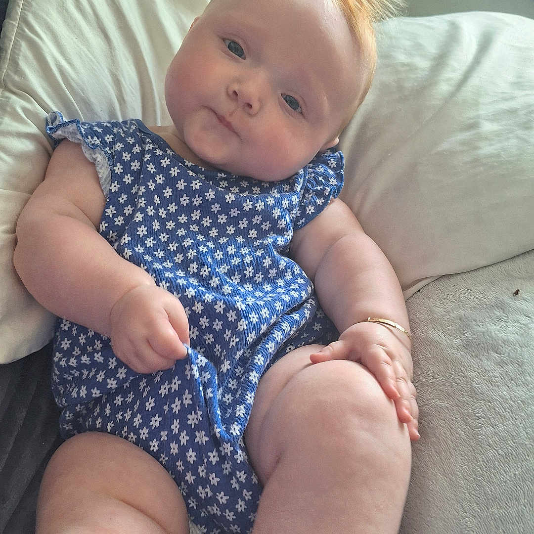 Delilah Denise is registered to the contest to win money with this photo: arms, baby, blue_outfit, child, chubby, couch, curious_expression, cute, floral_pattern, hand, indoors, infant, legs, pillow, plush, portrait, red_hair, relaxed, skin, soft_texture