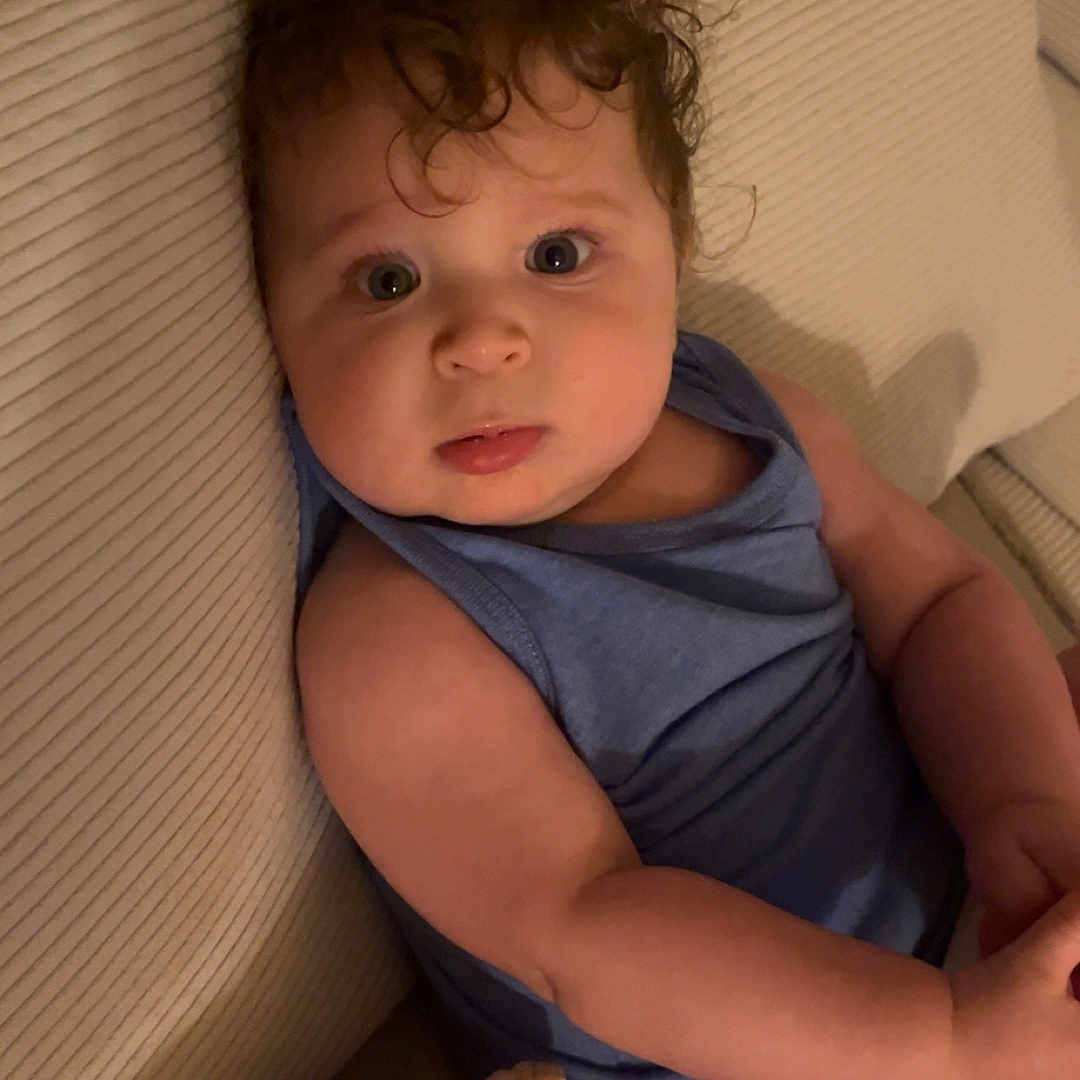 Noah participe au concours pour gagner de l'argent avec cette photo : arm, baby, beige, blanket, blue_top, casual, child, comfort, couch, curly_hair, diaper, expression, eyes, face, indoor, infant, person, seated, skin, texture