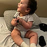 Kymari Briggs joined the competition — help win amazing prizes! baby, child, infant, onesie, striped_clothing, socks, bed, pillow, smiling, happy, curly_hair, indoor, soft_lighting, holding_object, black_sock, white_sock, cute, joyful, seated, looking_up