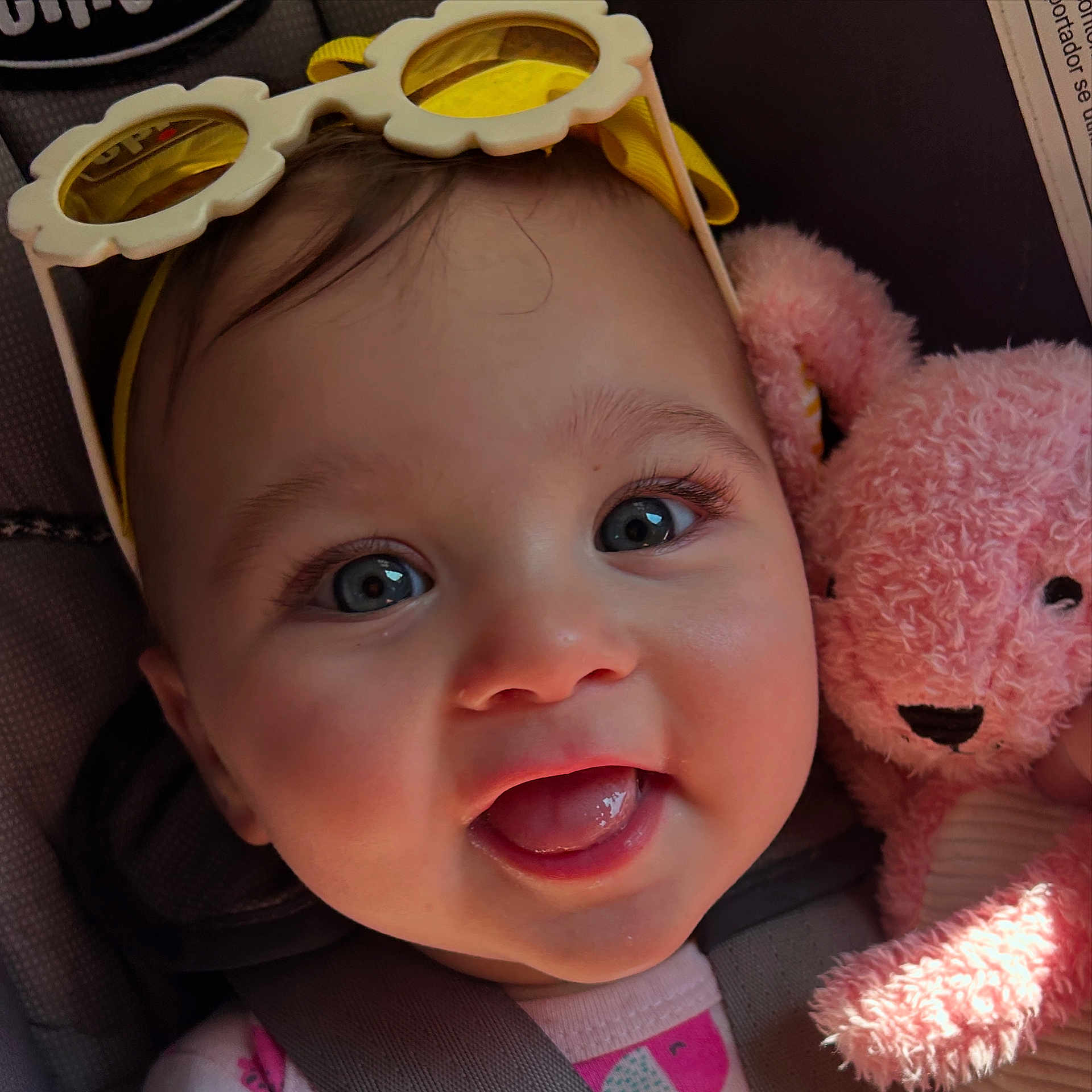 Eliza-Stylez is registered to the contest to win money with this photo: baby, child, smiling, face, blue_eyes, sunglasses, yellow, pink, teddy_bear, plush_toy, car_seat, seatbelt, closeup, cute, happy, infant, person, indoors, portrait, headwear