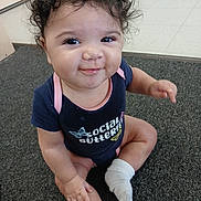 Leila is registered to the contest to win money with this photo: baby, child, curly_hair, smiling, onesie, socks, carpet, indoor, cute, person, infant, happy, seated, toddler, face, young_child, playful, skin, expression, clothing