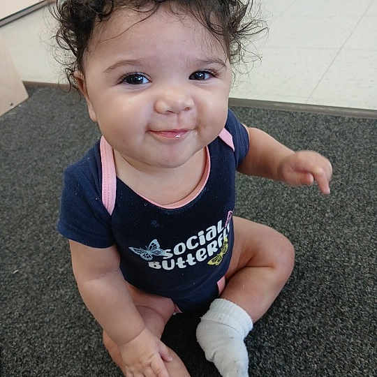 Leila is registered to the contest to win money with this photo: baby, child, curly_hair, smiling, onesie, socks, carpet, indoor, cute, person, infant, happy, seated, toddler, face, young_child, playful, skin, expression, clothing