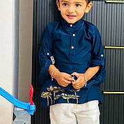 Yuvaan is registered to the contest to win money with this photo: child, toddler, boy, portrait, standing, smiling, happy, indoors, traditional_clothing, kurta, navy_blue, white_trousers, bracelets, red_mark, toy_racetrack, toy, playroom, door, fashion, full_body