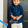 child, toddler, boy, portrait, standing, smiling, happy, indoors, traditional_clothing, kurta, navy_blue, white_trousers, bracelets, red_mark, toy_racetrack, toy, playroom, door, fashion, full_body