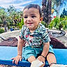 child, toddler, baby, boy, smiling, portrait, outdoor, garden, palm_trees, tropical, blue_sky, bench, seated, clothing, patterned_shirt, shorts, sneakers, bracelet, playground, sunny