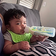 Gabriel is registered to the contest to win money with this photo: baby, bottle, feeding, green_bib, couch, cushion, indoor, window_blinds, hand, milk, pattern, resting, child, person, skin, face, short_hair, casual_clothing, comfort, nursing