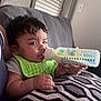 baby, bottle, feeding, green_bib, couch, cushion, indoor, window_blinds, hand, milk, pattern, resting, child, person, skin, face, short_hair, casual_clothing, comfort, nursing