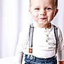 child, toddler, portrait, smile, suspenders, white_shirt, blue_pants, indoor, studio, curtains, hands, eyes, blond_hair, cute, fashion, buttoned_shirt, sitting, closeup, soft_lighting, high_key