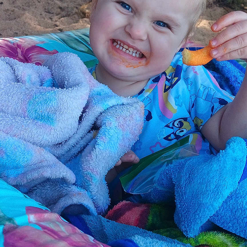 Scarlette is registered to the contest to win money with this photo: child, toddler, smiling, blanket, snack, outdoor, nature, trees, dirt, colorful, happy, person, face, clothing, playful, young_child, expression, cozy, relaxed, daytime