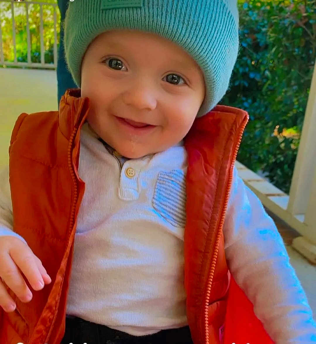 Kashton is registered to the contest to win money with this photo: toddler, child, smiling, hat, vest, outdoor, porch, greenery, happy, cute, face, clothing, buttoned_shirt, warm_clothes, baby, person, nature, daylight, casual, portrait