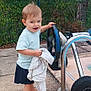 toddler, child, barefoot, steering_wheel, blanket, outdoor, fence, greenery, pavement, metal_frame, wheel, happy, smiling, shorts, tshirt, person, daylight, nature, tree, playground