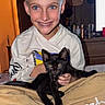 bed, black_cat, blue_eyes, casual_clothing, child, collar, furniture, happy, holding, hoodie, indoor, kitten, lamp, pants, person, pet, relaxed, room, smiling, young