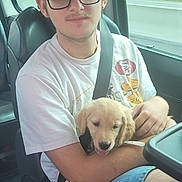 Baīa a rejoint le concours — aidez-le/la à gagner de superbes lots ! young_man, glasses, black_cap, white_tshirt, blue_shorts, puppy, golden_retriever, dog, car_interior, car_seat, seatbelt, window, trees, road, daytime, casual, smiling, pet, holding, relaxed