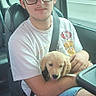 young_man, glasses, black_cap, white_tshirt, blue_shorts, puppy, golden_retriever, dog, car_interior, car_seat, seatbelt, window, trees, road, daytime, casual, smiling, pet, holding, relaxed