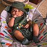 baby, baby_accessory, baby_seat, carpet, child, colorful_pattern, comfortable, cute, green_clothing, hand_in_mouth, hat, indoor, infant, infant_fashion, playful, portrait, relaxed, smiling, socks, young_child