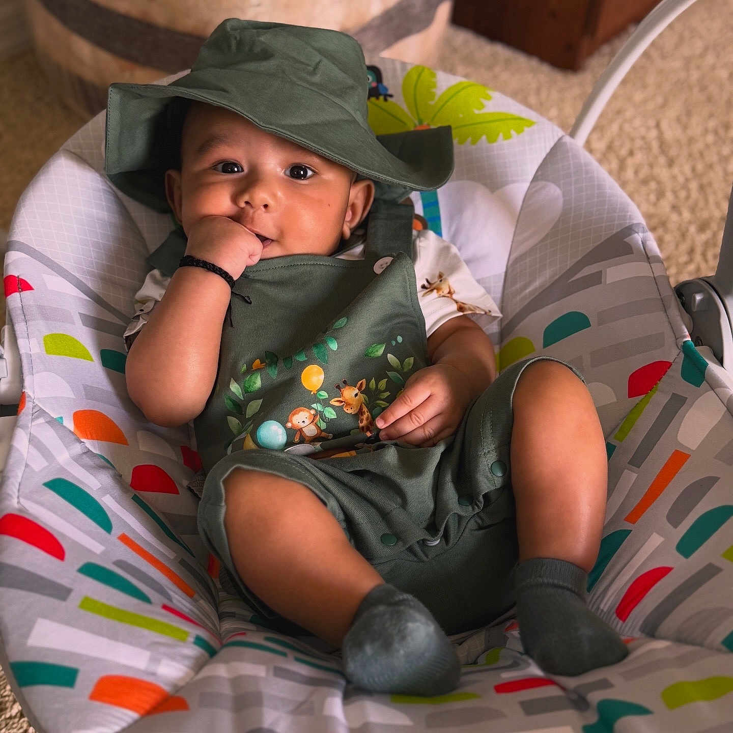 Vivaan is registered to the contest to win money with this photo: baby, baby_accessory, baby_seat, carpet, child, colorful_pattern, comfortable, cute, green_clothing, hand_in_mouth, hat, indoor, infant, infant_fashion, playful, portrait, relaxed, smiling, socks, young_child