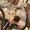 toddler, child, bed, pajamas, blanket, pillow, nightstand, water_bottle, baby, curly_hair, indoor, furniture, bedroom, table, clothing, person, reaching, cozy, curious, home