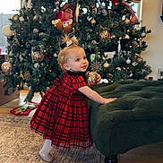 Aurora is registered to the contest to win money with this photo: toddler, child, christmas_tree, ornament, red_dress, plaid, holiday, decorations, indoor, carpet, green_ottoman, wood_floor, smiling, cute, festive, lights, toy, hair_tie, happy, seasonal