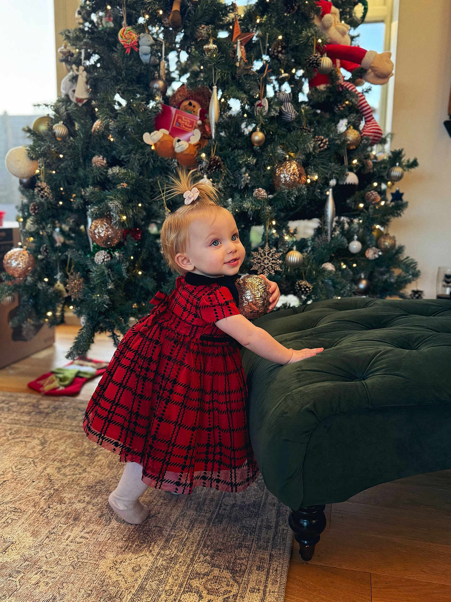 Aurora is registered to the contest to win money with this photo: toddler, child, christmas_tree, ornament, red_dress, plaid, holiday, decorations, indoor, carpet, green_ottoman, wood_floor, smiling, cute, festive, lights, toy, hair_tie, happy, seasonal