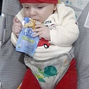Hudson is registered to the contest to win money with this photo: baby, child, red_hat, red_pants, baby_seat, gerber_food, food_pouch, infant, cute, chewing, indoor, colorful_background, smiling_sun, monkey_illustration, clothing, seat_belt, toddler, person, face, hands