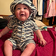 Waylon is registered to the contest to win money with this photo: baby, child, zebra_striped_clothing, hat, smile, sitting, bed, red_bedspread, indoor, clothes_hanging, happy, cute, chubby_cheeks, person, infant, shorts, buttoned_shirt, room, pillow, soft_toy