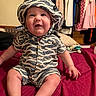 baby, child, zebra_striped_clothing, hat, smile, sitting, bed, red_bedspread, indoor, clothes_hanging, happy, cute, chubby_cheeks, person, infant, shorts, buttoned_shirt, room, pillow, soft_toy