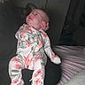 baby, infant, child, floral_onesie, patterned_clothing, sitting, supported, face, portrait, indoor, bedroom, bed, blanket, pillow, adult_hand, head_support, flash_photography, dim_lighting, candid, family_photo