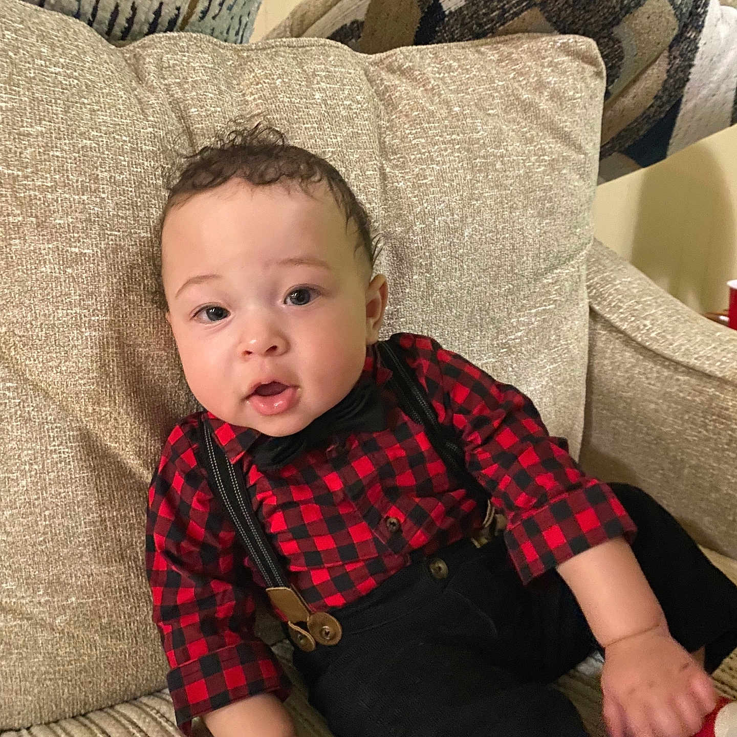 Zyaire is registered to the contest to win money with this photo: baby, infant, child, couch, clothing, red_and_black_checkered_shirt, suspenders, bow_tie, curly_hair, indoor, furniture, pillow, sitting, hand, face, portrait, cute, expression, young_child, comfort