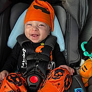Zyaire is registered to the contest to win money with this photo: baby, child, car_seat, halloween, orange_hat, orange_pants, black_shirt, smiling, happy, infant, seatbelt, plush_toy, toy, black_straps, cute, portrait, indoors, clothing, sitting, headwear