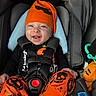 baby, child, car_seat, halloween, orange_hat, orange_pants, black_shirt, smiling, happy, infant, seatbelt, plush_toy, toy, black_straps, cute, portrait, indoors, clothing, sitting, headwear
