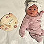 adorable, baby, bow, cozy, flower_decor, headband, infant, knitted_outfit, looking_up, lying_down, milestone_month, milestone_sign, newborn, pink_sweater, portrait_photo, soft_texture, tiny_hand, white_blanket, wide_eyes, wooden_prop