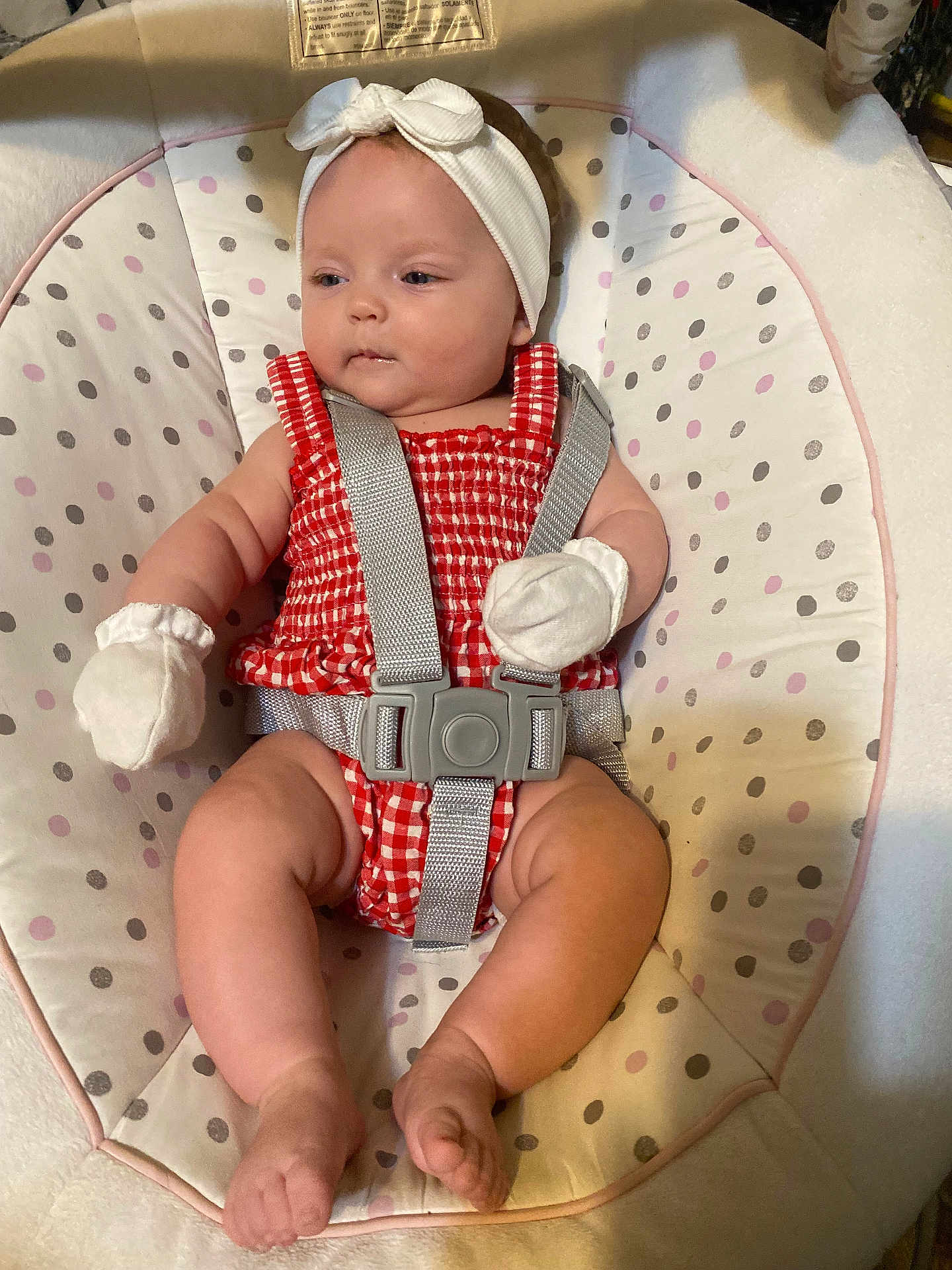 Ellie is registered to the contest to win money with this photo: baby, infant, baby_seat, headband, mittens, red_outfit, checkered_pattern, cushion, straps, sitting, barefoot, soft_lighting, indoors, cute, child, person, comfort, relaxed, young, adorable