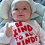 baby, infant, smiling, pink_clothing, text_on_clothing, cushion, seat, hand, face, cheerful, indoors, child, cute, gesture, happy, young, person, closeup, apparel, message