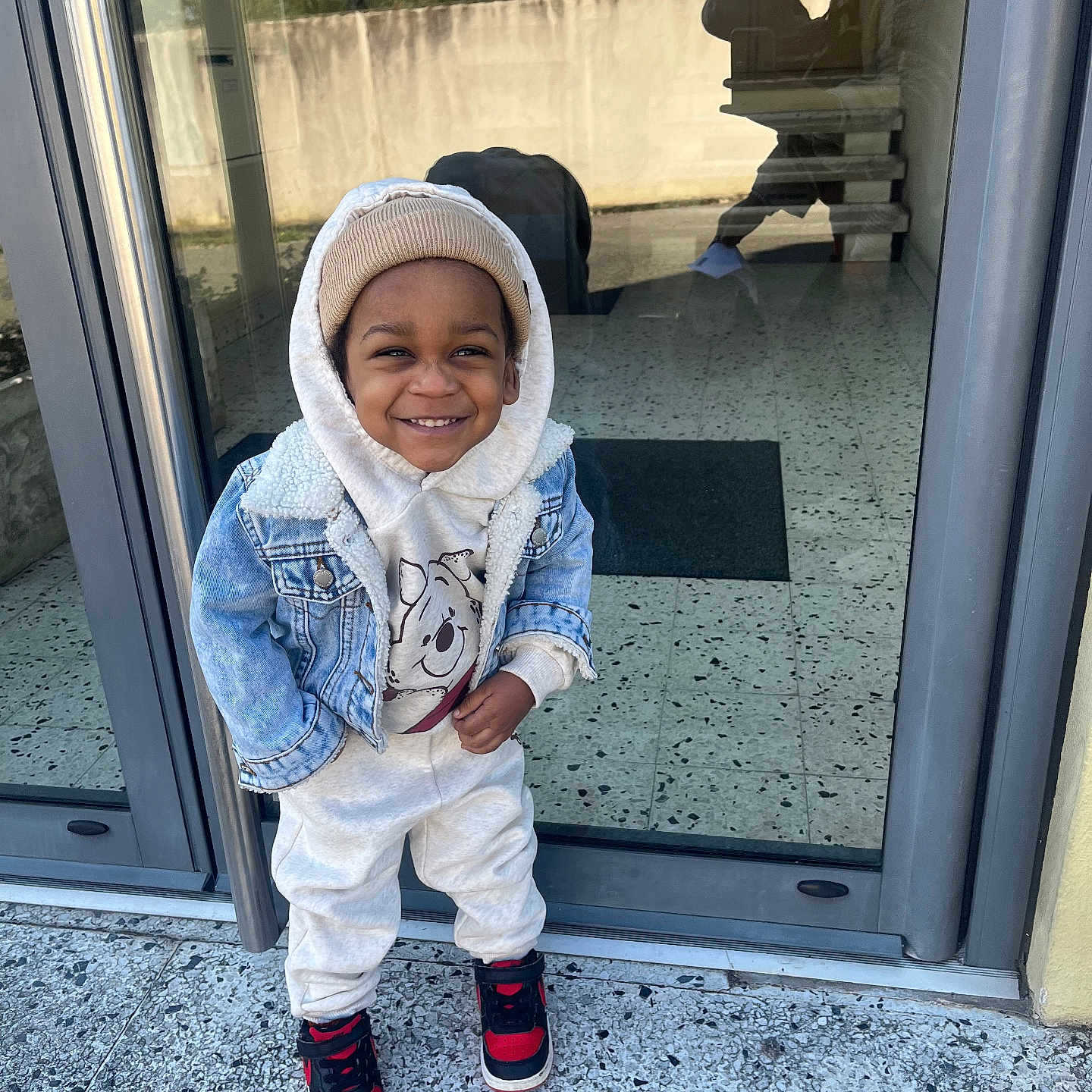 Tyler-Isaac participe au concours pour gagner de l'argent avec cette photo : beanie, casual_clothing, child, concrete, denim_jacket, floor, glass_door, happy, hoodie, outdoor, person, portrait, reflection, sidewalk, smiling, sneakers, standing, toddler, urban, winter_wear