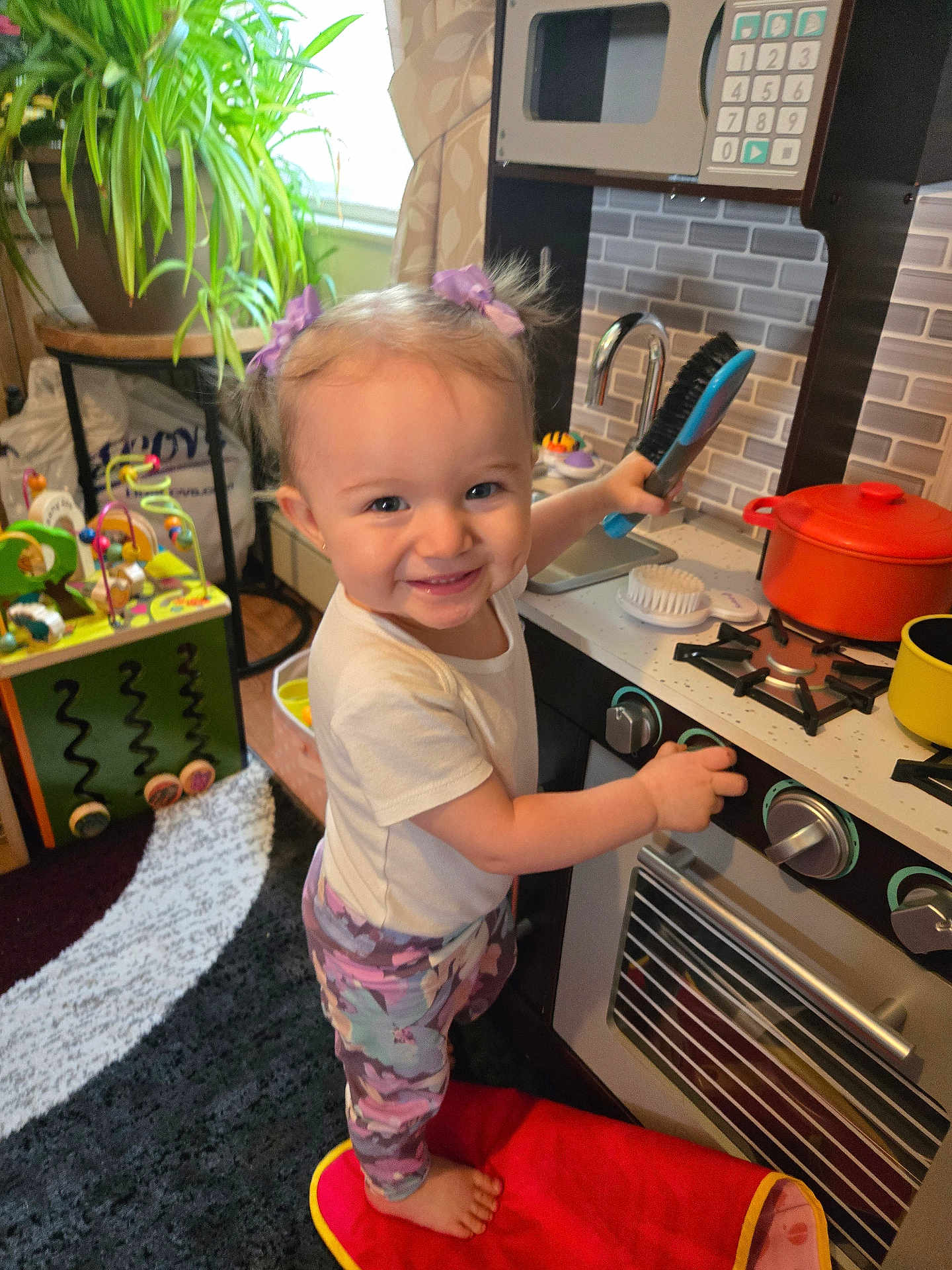 Kailyana joined the competition — help win amazing prizes! toddler, child, toy_kitchen, play, smile, indoor, hair_bows, barefoot, cushion, toy_pot, toy_brush, carpet, plant, furniture, window, curtain, colorful, happy, pretend_play, living_room