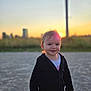 child, toddler, sunset, outdoor, smiling, jacket, face, person, nature, cityscape, sunlight, sky, grass, evening, portrait, young_child, casual_clothing, happy, warm_light, standing