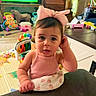 baby, blanket, cartoon_tv, child, colorful, couch, curious, cute, expression, floor, furniture, home, indoor, living_room, person, pink_bow, pink_tank_top, play_area, toys, young_child