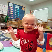 Hailey is registered to the contest to win money with this photo: baby, child, toddler, smiling, happy, waving, portrait, red_shirt, classroom, daycare, playroom, toys, colorful, indoor, crib, play_mat, infant, standing, blond_hair, photograph