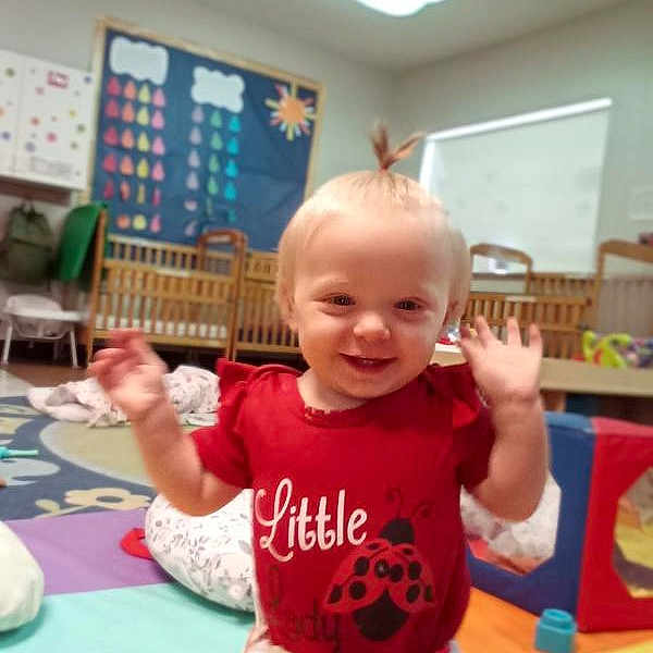 Hailey is registered to the contest to win money with this photo: baby, child, toddler, smiling, happy, waving, portrait, red_shirt, classroom, daycare, playroom, toys, colorful, indoor, crib, play_mat, infant, standing, blond_hair, photograph