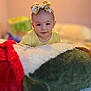 baby, bed, blanket, bow, child, cozy, cute, face, focused, headband, indoor, lying_down, person, plush_toy, portrait, smiling, soft_toy, warm_lighting, yellow_clothing, young_child