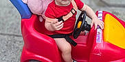 Tinatin is registered to the contest to win money with this photo: toddler, child, toy_car, red_clothing, red_hat, plush_toy, sidewalk, steering_wheel, car_toy, blue_wheels, outdoor, person, concrete, street, seatbelt, pink_plush, footwear, expression, vehicle_wheel, urban