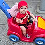 toddler, child, toy_car, red_clothing, red_hat, plush_toy, sidewalk, steering_wheel, car_toy, blue_wheels, outdoor, person, concrete, street, seatbelt, pink_plush, footwear, expression, vehicle_wheel, urban