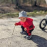 bicycle_wheel, bucket_hat, candid, child, curious, exploring, gravel_ground, outdoors, park, person, play, portrait, red_hoodie, shadow, sneakers, squatting, stick, sunlight, toddler, trees